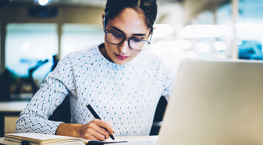 En kvinna sitter vid datorn och skriver samtidigt i ett block.